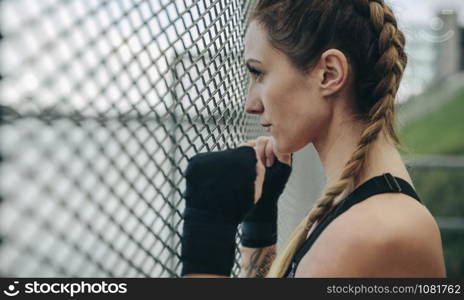 Young sportswoman with boxing bandages looking through a fence outdoors. Sportswoman looking through a fence