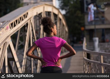 Young sporty african american woman running on sidewalk across the bridge at early morning jogging with city sunrise scene in background