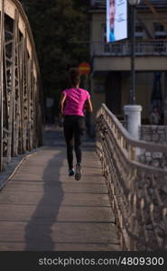 Young sporty african american woman running on sidewalk across the bridge at early morning jogging with city sunrise scene in background