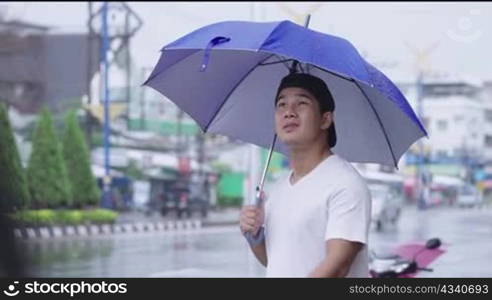 Young sporty asian man holding umbrella standing on the street side on the rainy day, unsatisfied stuck in the rain, unlucky day while going out, Asia tropical climate season changes, pouring rain