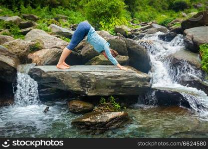 Young sporty fit woman doing yoga asana Adho mukha svanasana - downward facing dog - at tropical waterfall. Woman doing yoga oudoors at tropical waterfall