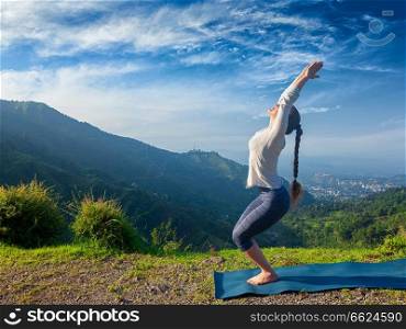 Young sporty fit woman doing yoga asana Utkatasana (chair pose) outdoors in mountains Himalayas in the morning. Himachal Pradesh, India. Woman doing yoga asana Utkatasana outdoors