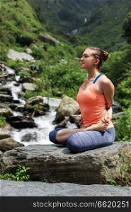 Young sporty fit woman doing yoga - meditating in Baddha Padmasana (Bound Lotus Pose) outdoors at tropical waterfall. Young woman does yoga oudoors at waterfall