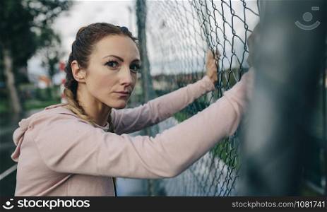 Young sporty girl with challenging look resting on sports court fence. Sporty girl resting on sports court fence