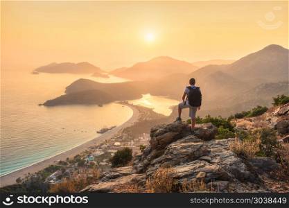 Young sporty man with backpack standing on the top of rock and looking at the seashore and mountains at sunset in summer. Scene with man, sea, mountain ridges and orange sky with sun. Oludeniz, Turkey. Young sporty man with backpack, seashore and mountains