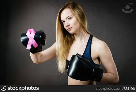 Young sporty woman wearing boxing gloves having pink ribbon tape, breast cancer symbol. Fighting with disease, feminine motivation. Studio shot on dark background.. Young woman wearing boxing gloves having pink ribbon