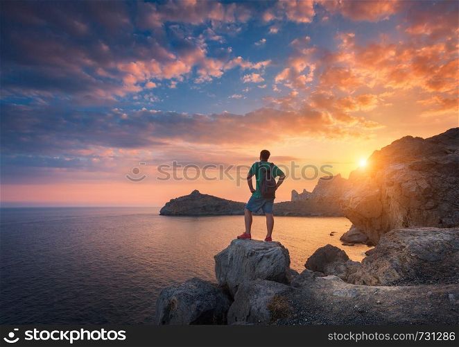 Young standing man with backpack on the stone on the seashore at colorful sunset sky. Beautiful landscape with sporty man, rocks, sea and clouds at sunset. Sport, lifestyle background. Travel.