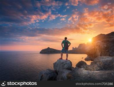 Young standing man with backpack on the stone on the seashore at colorful sunset sky. Beautiful landscape with sporty man, rocks, sea and clouds at sunset. Sport, lifestyle background. Travel.