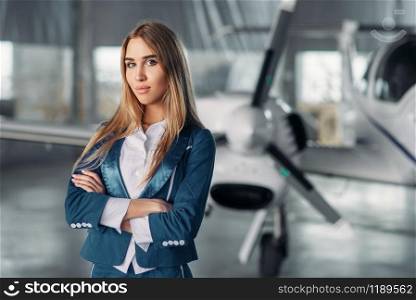 Young stewardess in uniform poses against propeller plane in hangar. Air hostess in suit near airplane. Private airline, flight attendant. Stewardess against propeller plane in hangar