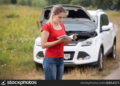 Young stressed woman standing at broken car and reading owner manual