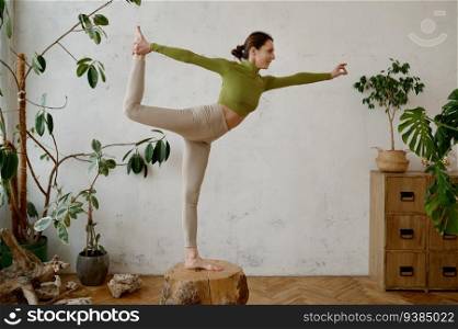 Young strong woman balancing on wooden stump in dancer yoga pose Natarajasana. Active body training at home. Young strong woman balancing on wooden stump in dancer yoga pose Natarajasana