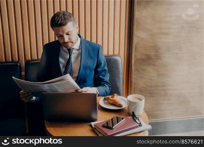 Young stubbly man entrepreneur in formal wear focused on reading financial chronicle in newspaper during coffee break while enjoying his cappuccino and croissant, sitting at table with open laptop. Young man entrepreneur in formal tuxedo reading newspaper in cafe
