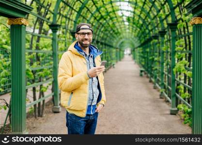 Young student male with beard having stylish clothes standing against green background using mobile phone for downloading video. Bearded hipster in yellow anorak resting outdoors typing messages
