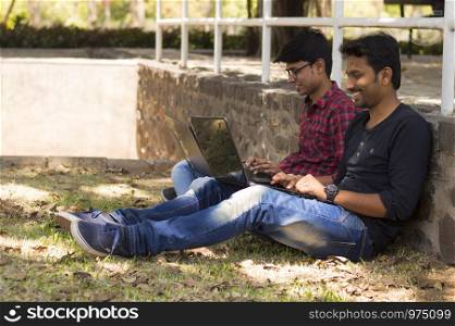 Young students using laptop lean on wall beside each other in park
