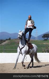 Young stunt man in casual outfit riding white horse on sandy ground