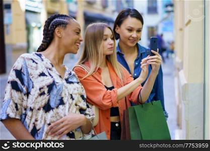 Young stylish lady taking photo pf fashion boutique showcase on smartphone during shopping with diverse female friends in city. Fashionable multiracial ladies taking photo on smartphone during shopping in city