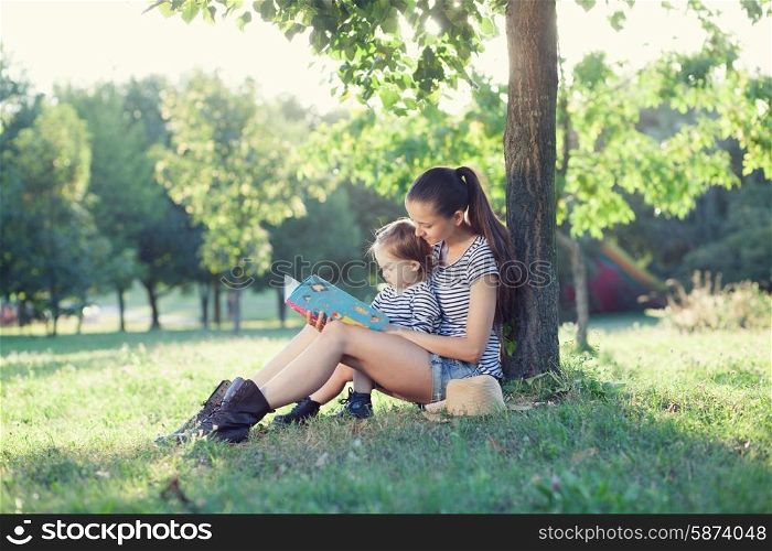 Young stylish mother and toddler reading book at garden during summer fun