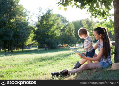 Young stylish mother and toddler reading book at garden during summer fun