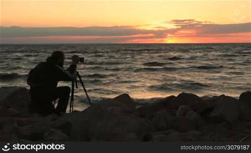Young stylish photographer with backpack making photos of sea and rocks with camera on a tripod in twilight time. Professional photographer shooting seascape in glow of beautiful red sunset at seaside. Slow motion.