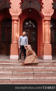 Young stylish pretty couple of hipsters in love are having fun in the city near castle in spring. young blond woman in red dress and man with beard are smiling on a date.. Young stylish pretty couple of hipsters in love are having fun in the city near castle in spring. young blond woman in red dress and man with beard are smiling on a date