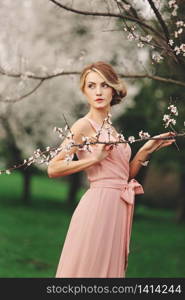 young stylish woman near blossoming flowering tree in the park. spring background. blonde girl with hairstyle in pink dress. copy space. young stylish woman near blossoming flowering tree in the park. spring background. blonde girl with hairstyle in pink dress. copy space.