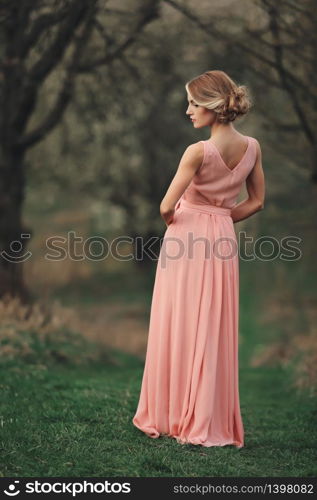 young stylish woman outdoors in the spring park. blonde girl with hairstyle in long pink dress possing. selective focus. back side view.. young stylish woman outdoors in the spring park. blonde girl with hairstyle in long pink dress possing. selective focus. back side view