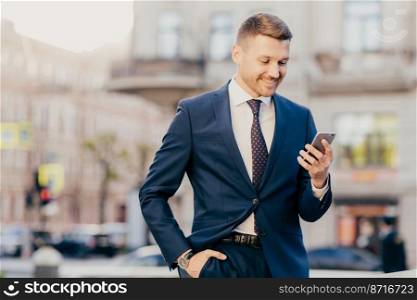 Young succesfful delighted happy businessman reads text message on modern cellular, wears elegant suit, holds hand in pocket, enjoys online communication. People, business, technology concept