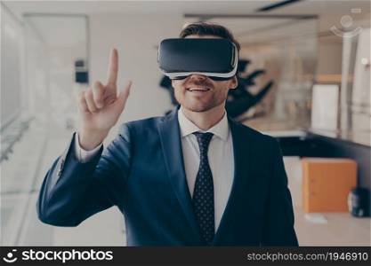 Young successful businessman in VR goggles touching air with finger during virtual business meeting, man in 3d glasses taking part in conference in augmented reality while working in office. Young successful businessman in VR goggles touching air with finger during virtual business meeting