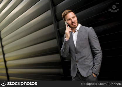 Young successful businessman wearing grey suit and holding his smartphone while standing near modern office or skyscrapers