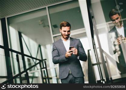 Young successful businessman wearing grey suit and holding his smartphone while standing near modern office or skyscrapers