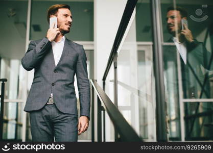 Young successful businessman wearing grey suit and holding his smartphone while standing near modern office or skyscrapers