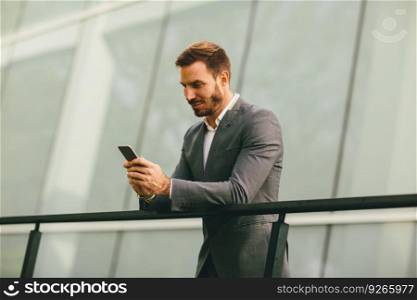 Young successful businessman wearing grey suit and holding his smartphone while standing near modern office or skyscrapers