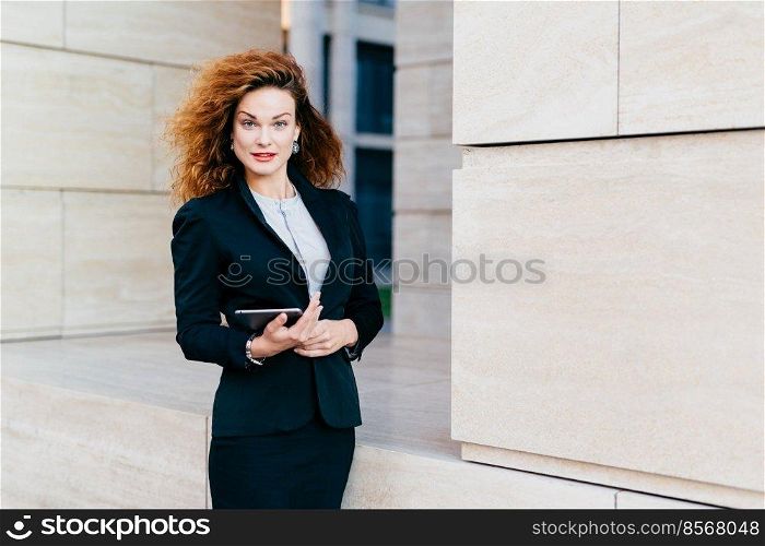 Young successful businesswoman in elegant clothes, having luxuriant hair, holding tablet in hands while going for work. Gorgeous female holding digital tablet computer. Technology and business