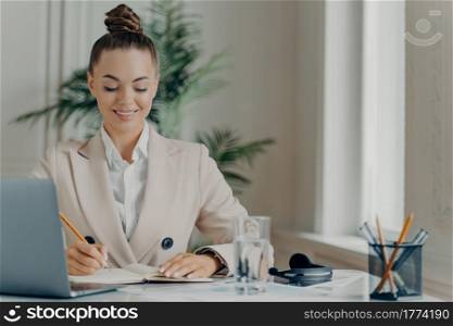 Young successful businesswoman in formal wear enjoying office work, writing down information in notepad with positive expression while sitting at workplace with laptop. Job and occupation concept. Young successful businesswoman in formal wear writing down information in notepad