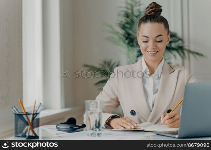 Young successful businesswoman in formal wear enjoying office work, writing down information in notepad with positive expression while sitting at workplace with laptop. Job and occupation concept. Young successful businesswoman in formal wear writing down information in notepad