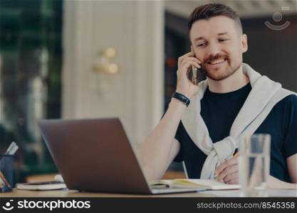 Young successful entrepreneur or male freelancer in casual wear talking on mobile phone and looking at laptop while sitting at his workplace in office or home, smiling and discussing business ideas. Happy businessman talking on mobile phone while working from home