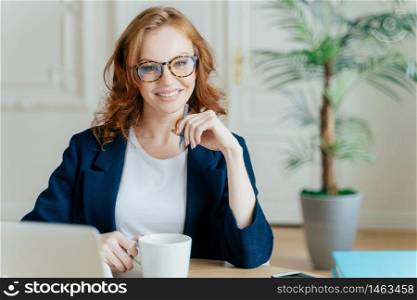 Young successful female entrepreneur prepares report for startup project, searches information on laptop computer, concentrated on her task, holds cup of coffee, pen keeps hand under chin poses indoor