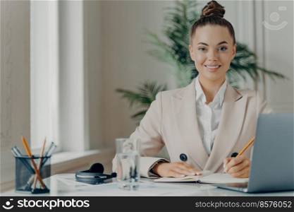 Young successful female freelancer enjoying remote work, positive businesswoman sitting at office desk and working on laptop computer, writing down information in notepad and smiling happily at camera. Young successful female freelancer writing down information in notepad