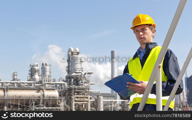 Young supervisor with a note board and pen in his hands, wearing a hard hat and safety vest in front of a petrochemical plant and refinery