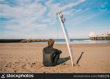 Young surfer sitting on sandy beach and next to his surfboard. Sport and water sport concept.