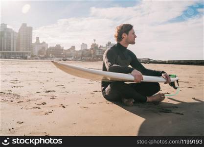 Young surfer sitting on sandy beach looking at the ocean with his surfboard. Sport and water sport concept.
