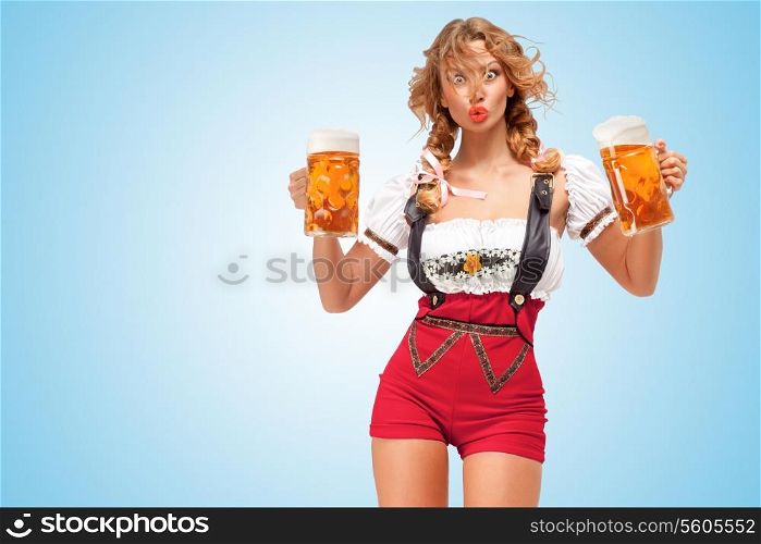 Young surprised sexy Swiss woman wearing red jumper shorts with suspenders in a form of a traditional dirndl, holding two beer mugs on blue background.