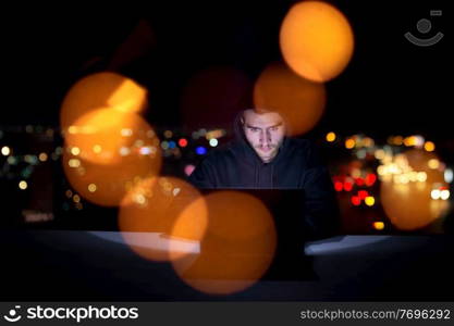 Young talented hacker using laptop computer while working in dark office with big city lights in the background at night