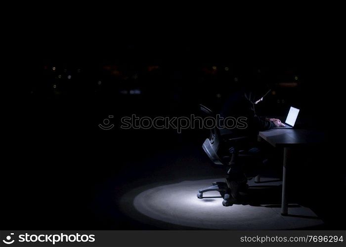 Young talented hacker using laptop computer while working in dark office with big city lights in the background at night