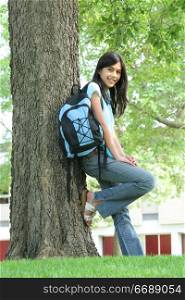 Young teen girl standing with backpack by tree, smiling. Part asian, Scandinavian descent.