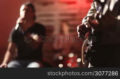 Young teenage rock band rehearsing in the garage. Focus on guitarist&acute;s hands picking chords on electric guitar then focus on frontman holding microphone and singing with drummer playing his kit on background while band practicing and training in club