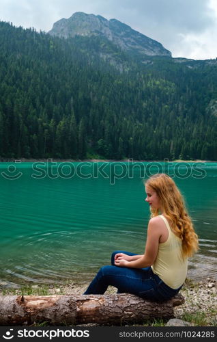 Young teenager girl rest on tree log on Black lake (Crno jezero) shore, summer landscape. Zabljak Municipality, Montenegro.
