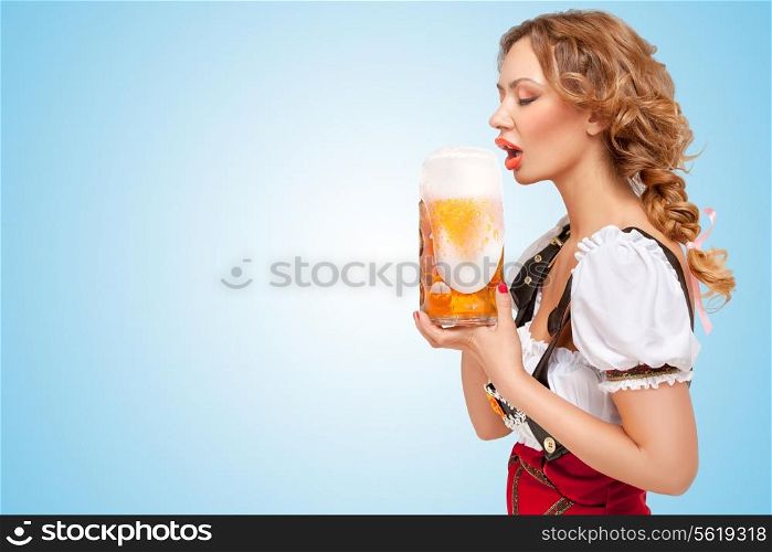 Young thirsty sexy Swiss woman wearing red jumper shorts with suspenders in a form of a traditional dirndl, drinking beer with froth flowing out the mug on blue background.