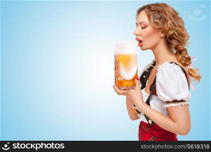 Young thirsty sexy Swiss woman wearing red jumper shorts with suspenders in a form of a traditional dirndl, drinking beer with froth flowing out the mug on blue background.