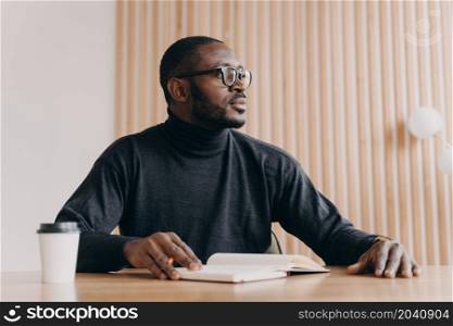Young thoughtful african american businessman sitting at desk with pen and agenda looking away with pensive expression. Millennial biracial entrepreneur coming up with new ideas at home office. Thoughtful african businessman sits at desk with pen and agenda looks away with pensive expression